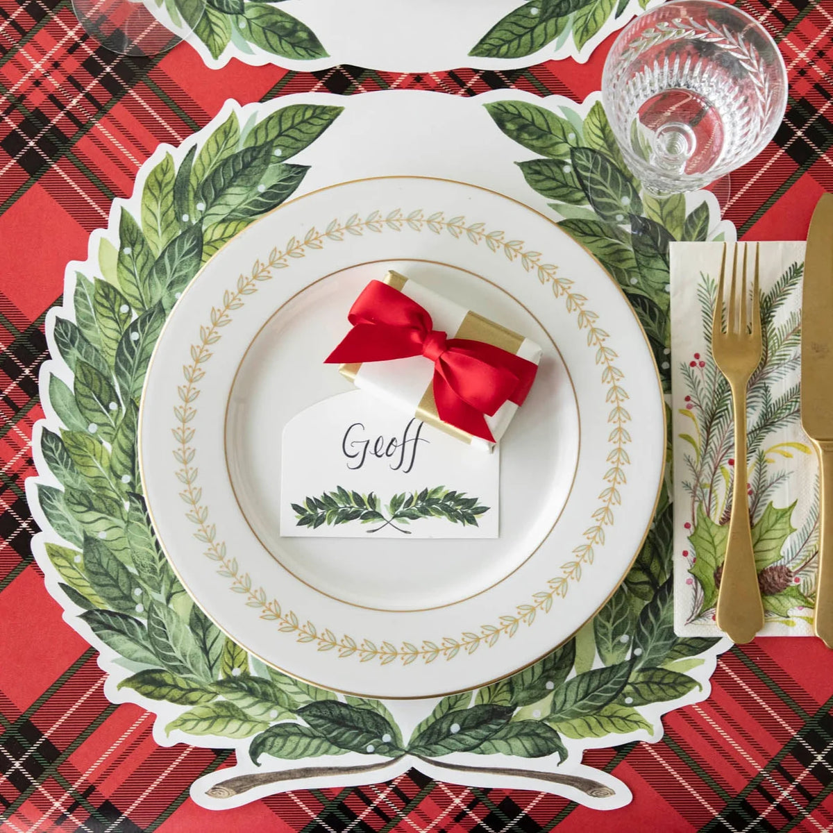 A festive holiday table setting featuring a white charger plate with a green holly leaf border, a red bow, and a place card reading 'Geoff' on a red plaid tablecloth.