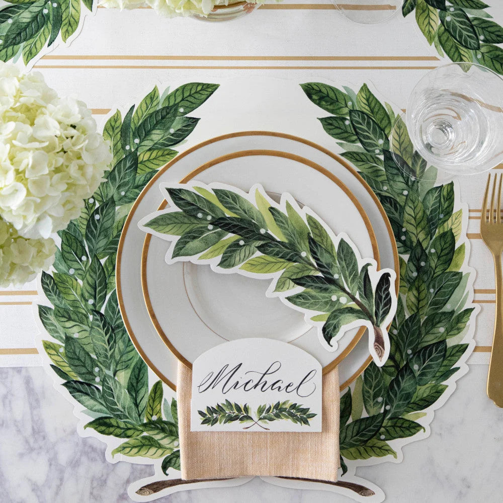 A styled table setting featuring a personalized place setting with a green leaf wreath placemat, a white plate with gold trim, a matching leaf-shaped napkin, a wooden name block reading 'Michael', and white flowers on a marble surface.