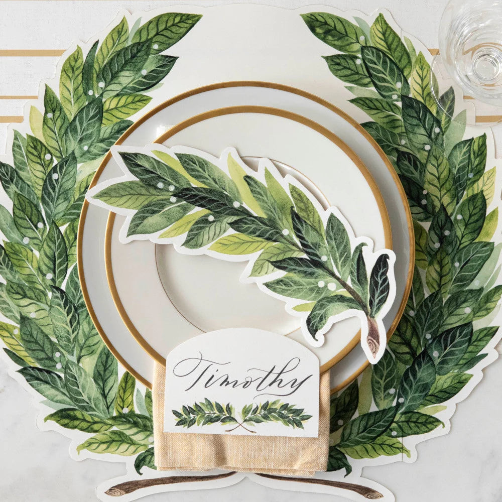 A styled table setting featuring a large green leaf wreath placemat with white plates, a green leaf-shaped napkin, and a personalized place card that reads 'Timothey'.