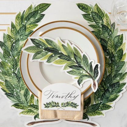 A styled table setting featuring a large green leaf wreath placemat with white plates, a green leaf-shaped napkin, and a personalized place card that reads 'Timothey'.