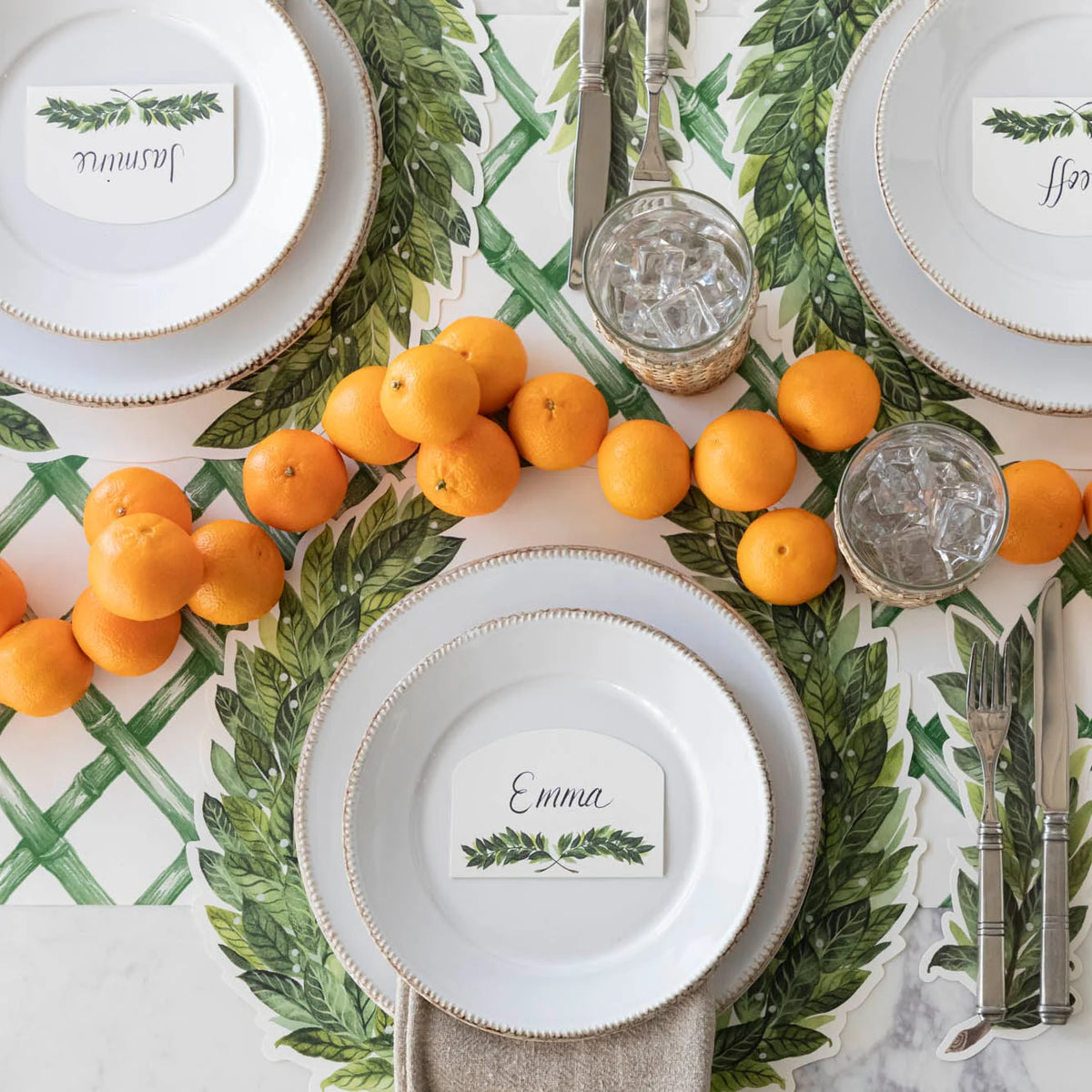 A styled table setting featuring a green and white botanical tablecloth with a laurel wreath pattern, white charger plates, clear glassware with ice, silver cutlery, and a green-and-white place card garland with oranges.