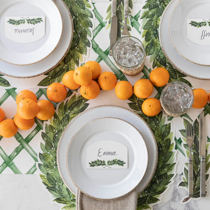 A styled table setting featuring a green and white botanical tablecloth with a laurel wreath pattern, white charger plates, clear glassware with ice, silver cutlery, and a green-and-white place card garland with oranges.
