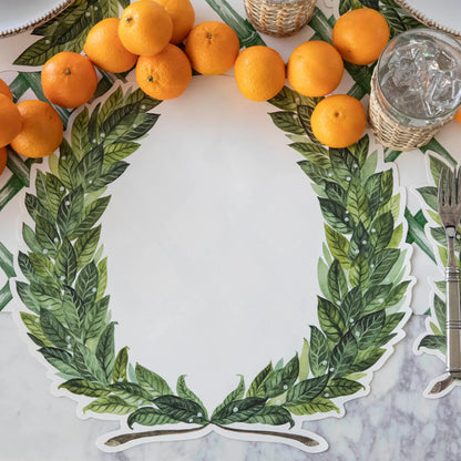 A circular table placemat with a green laurel wreath border and a blank center, styled on a marble surface with oranges around the edge and a glass of water nearby.