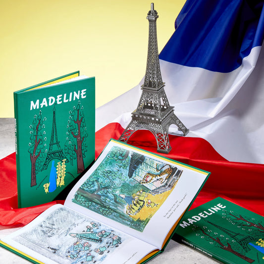 A children's book titled "Madeline" displayed open and closed, with a small metallic Eiffel Tower model and a French flag in the background. The book cover is green with illustrated trees and the Eiffel Tower, and the open pages show colorful illustrated scenes.