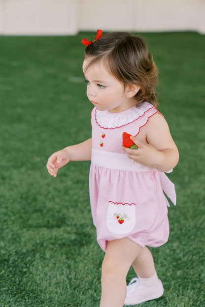 A pink baby bubble romper with white trim and strawberry appliques on the pockets and a ruffled collar.