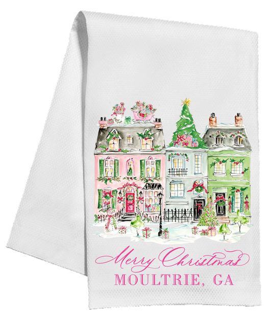 A white kitchen tea towel featuring a colorful watercolor illustration of holiday houses in Moultrie, GA, with 'Merry Christmas' and 'MOULTRIE, GA' printed in pink script and block lettering at the bottom.