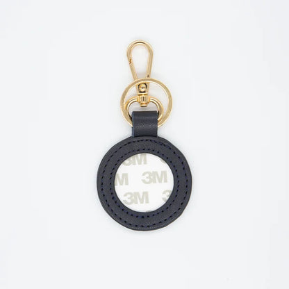 A black circular leather keychain tag with a gold-tone metal clasp and key ring, featuring a visible 3M adhesive backing on the inside of the ring, shot against a plain white background.