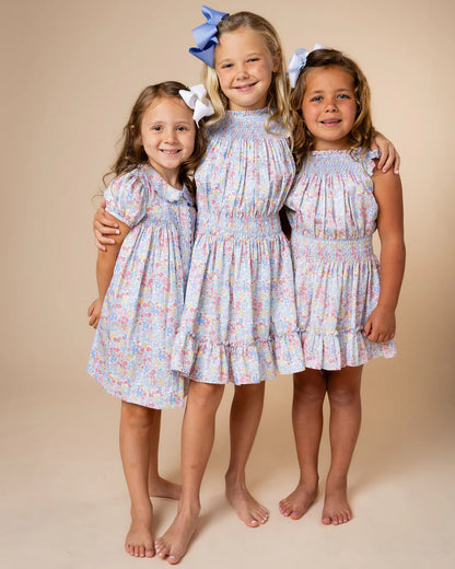 Three young girls standing together and smiling, wearing matching smocked dress dresses with a light blue, pink, and white floral print, set against a solid beige studio background.