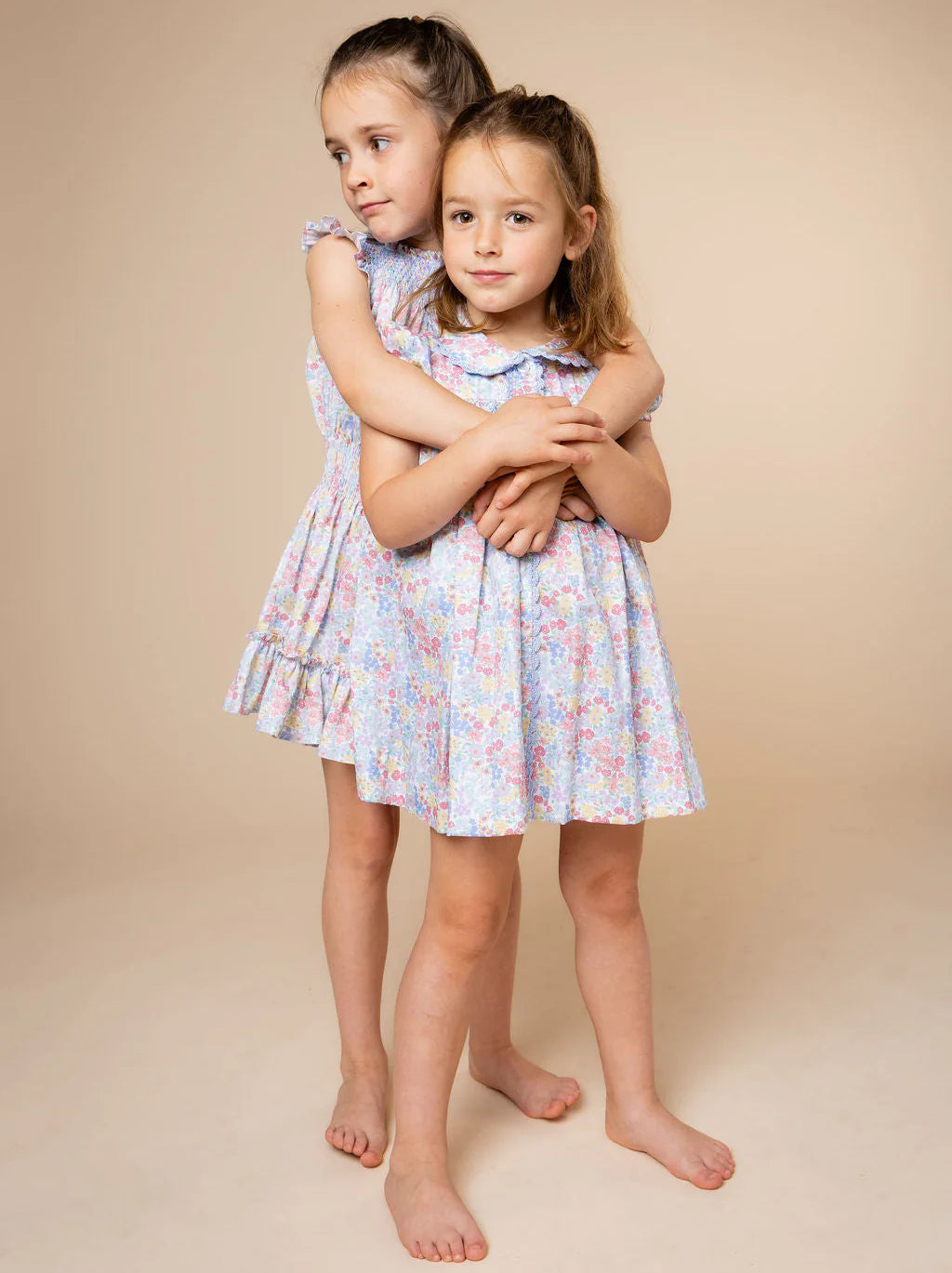 Two young girls standing barefoot against a plain beige studio background, wearing matching light blue floral dresses with pink and white details.