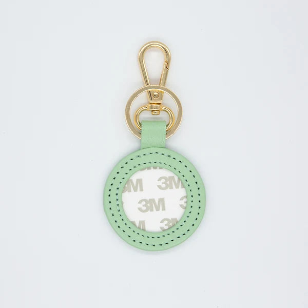 A mint green leather keychain or bag charm with a gold-tone swivel clasp and a circular cutout showing a white 3M adhesive backing, set against a plain white background.
