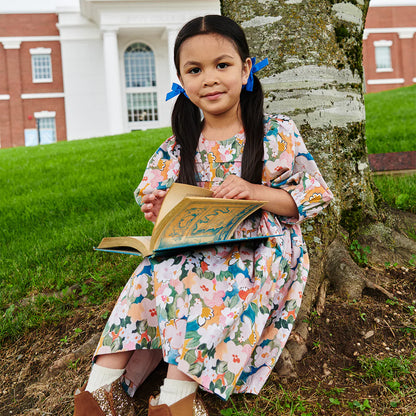 Young girl in a floral dress reading a book outdoors near a tree
