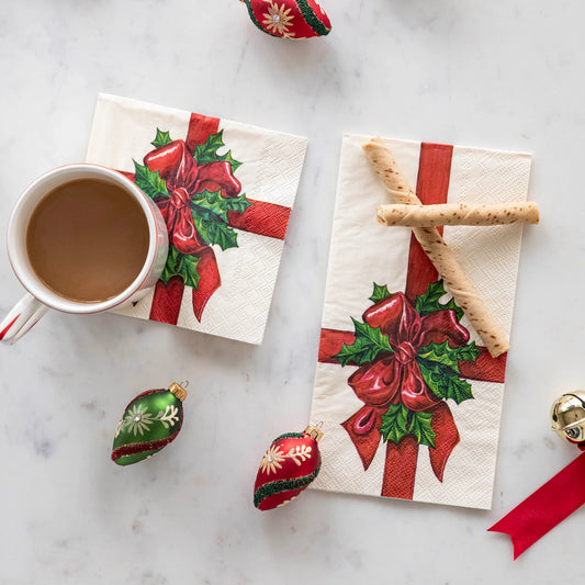 Napkins with Christmas designs featuring red bows and holly leaves, accompanied by a cup of coffee and decorative ornaments on a marble surface.