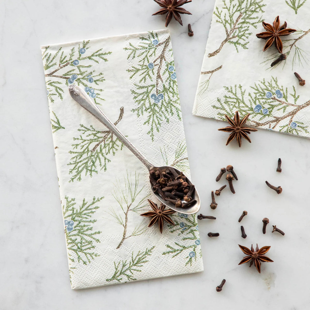 Two cream paper napkins with a green and blue pine branch pattern are arranged on a white marble surface, one with a silver spoon holding cloves and star anise scattered around.