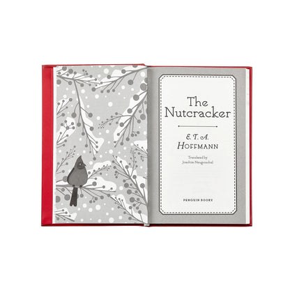 An open hardcover book with a red cover and gray-and-white illustrated interior pages. The left page shows a bird perched among winter branches, and the right page displays the title 'The Nutcracker & T.