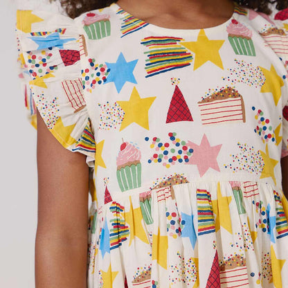 Child wearing a colorful dress with star, cake, and cupcake patterns on a plain background