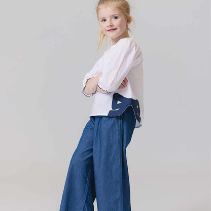 Young girl wearing a white blouse with a blue design and blue pants on a plain background