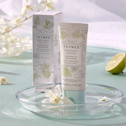 Thymes Bergamot Nectar hand cream and packaging on a light blue surface with flowers and a lime.
