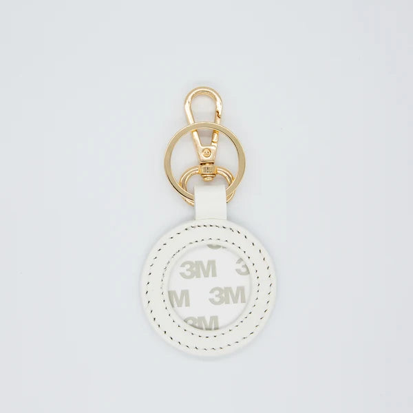 A white circular keychain or bag charm with a gold-tone metal clasp and key ring, featuring a white strap and a printed white-on-white circular insert with repeating '3M' branding.