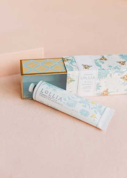 A tube of Lollia hand cream with floral design, next to a box with similar design, on a pink background.