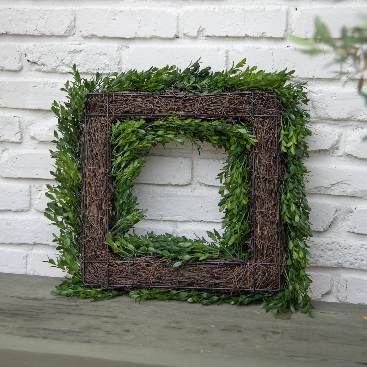 A square decorative wreath made of green boxwood leaves and brown woven vine, displayed against a white painted brick wall on a gray wooden surface.