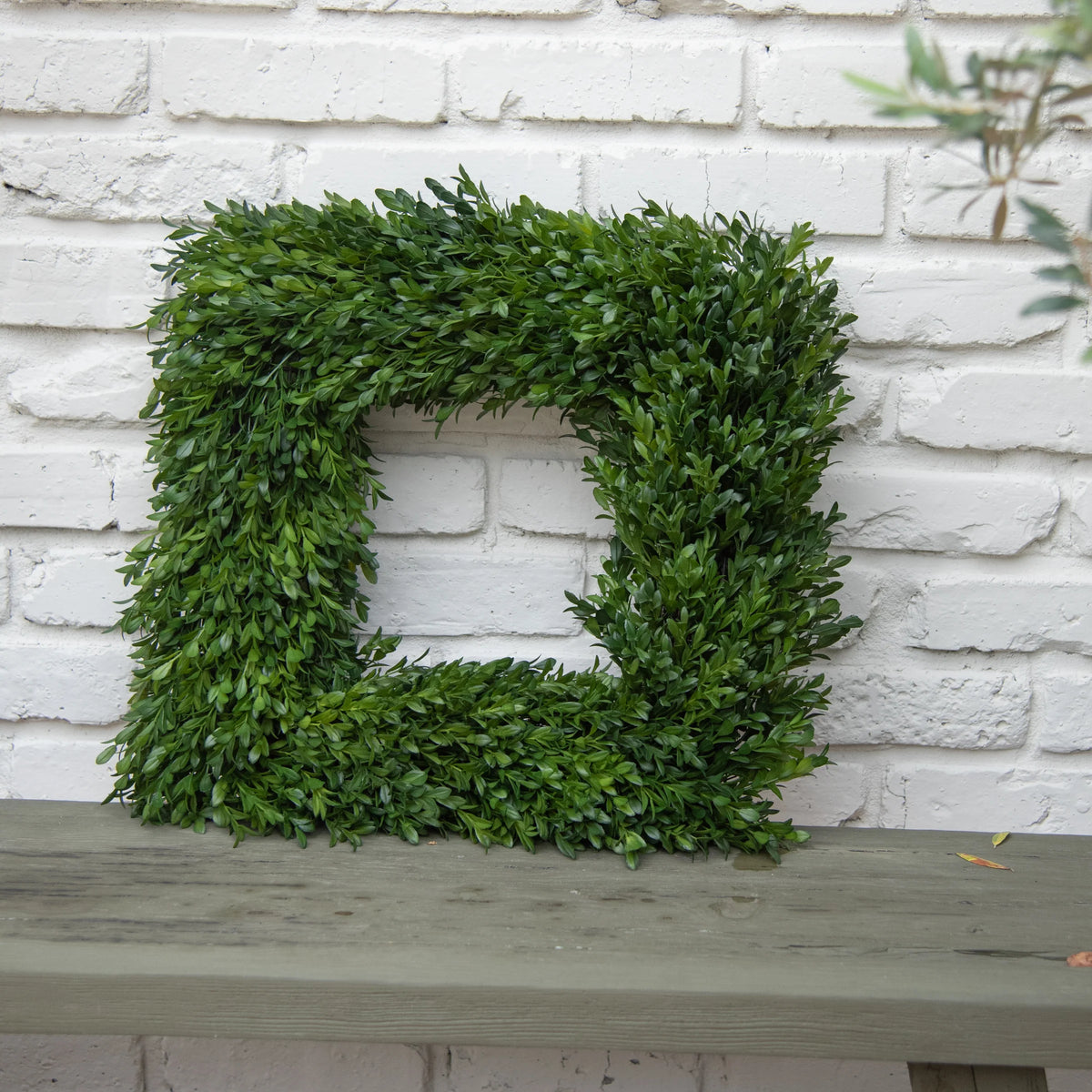 A square green boxwood wall wreath with a cutout center, displayed on a weathered gray wooden mantel against a white brick wall.