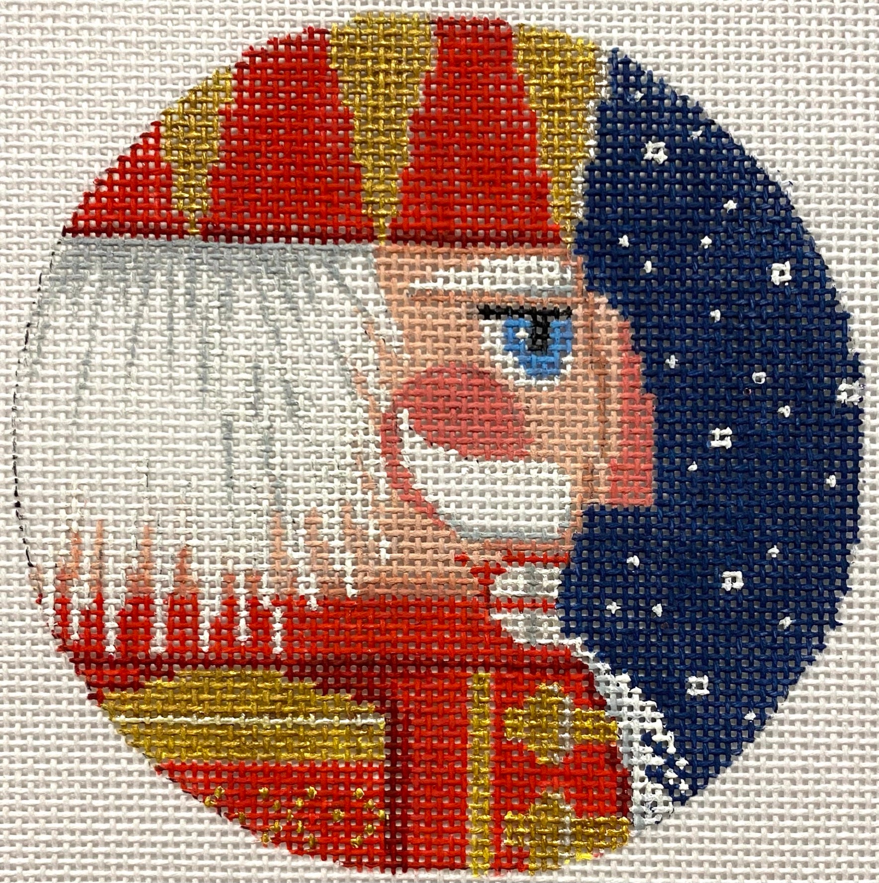 A circular needlepoint canvas featuring a stylized nutcracker profile. The nutcracker has a red and gold hat, white hair, a blue eye, a red coat, and is set against a dark blue starry background.