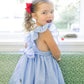 Young girl wearing a blue checkered dress with ruffled straps in a bright room.