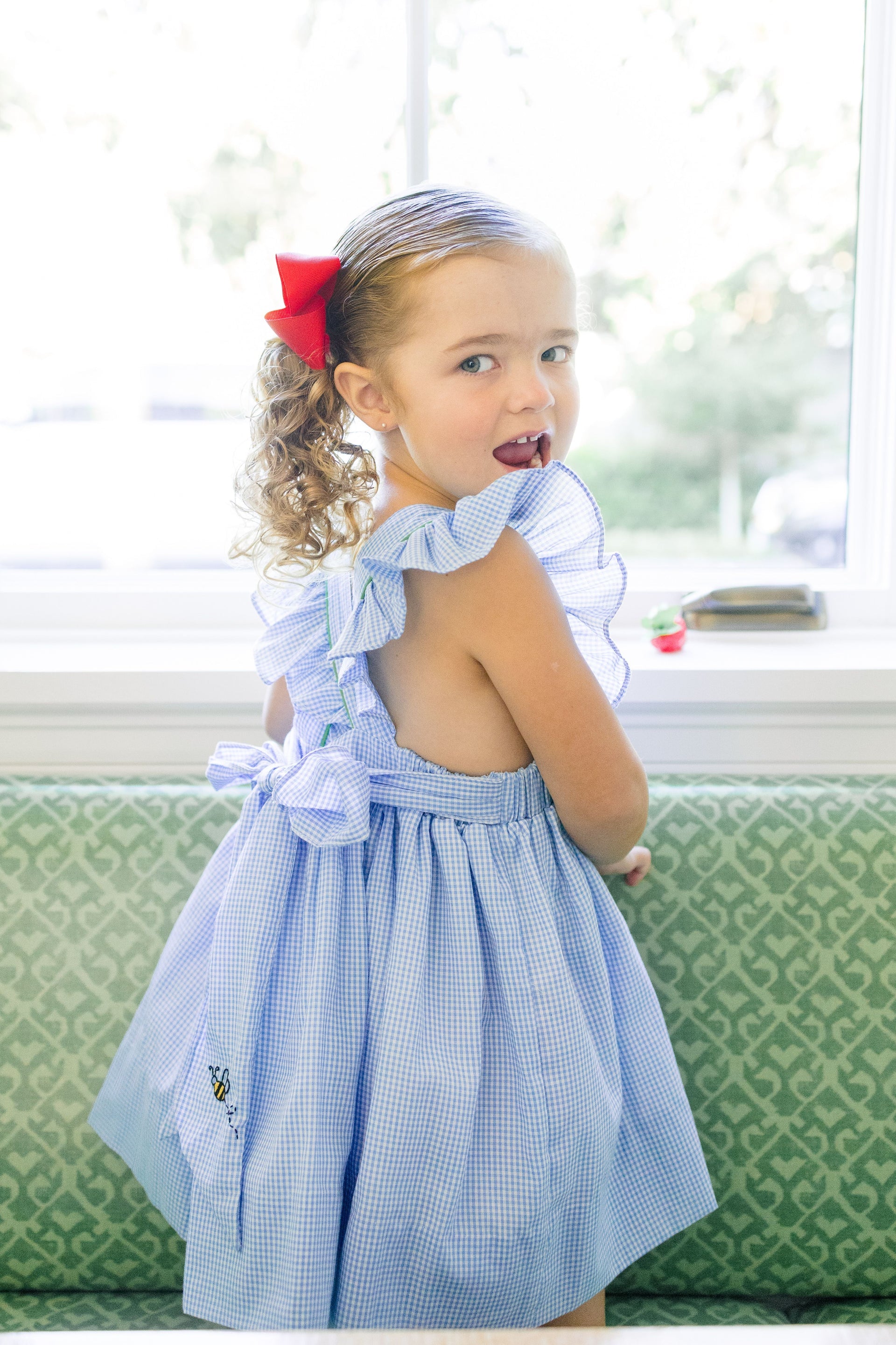 Young girl wearing a blue checkered dress with ruffled straps in a bright room.