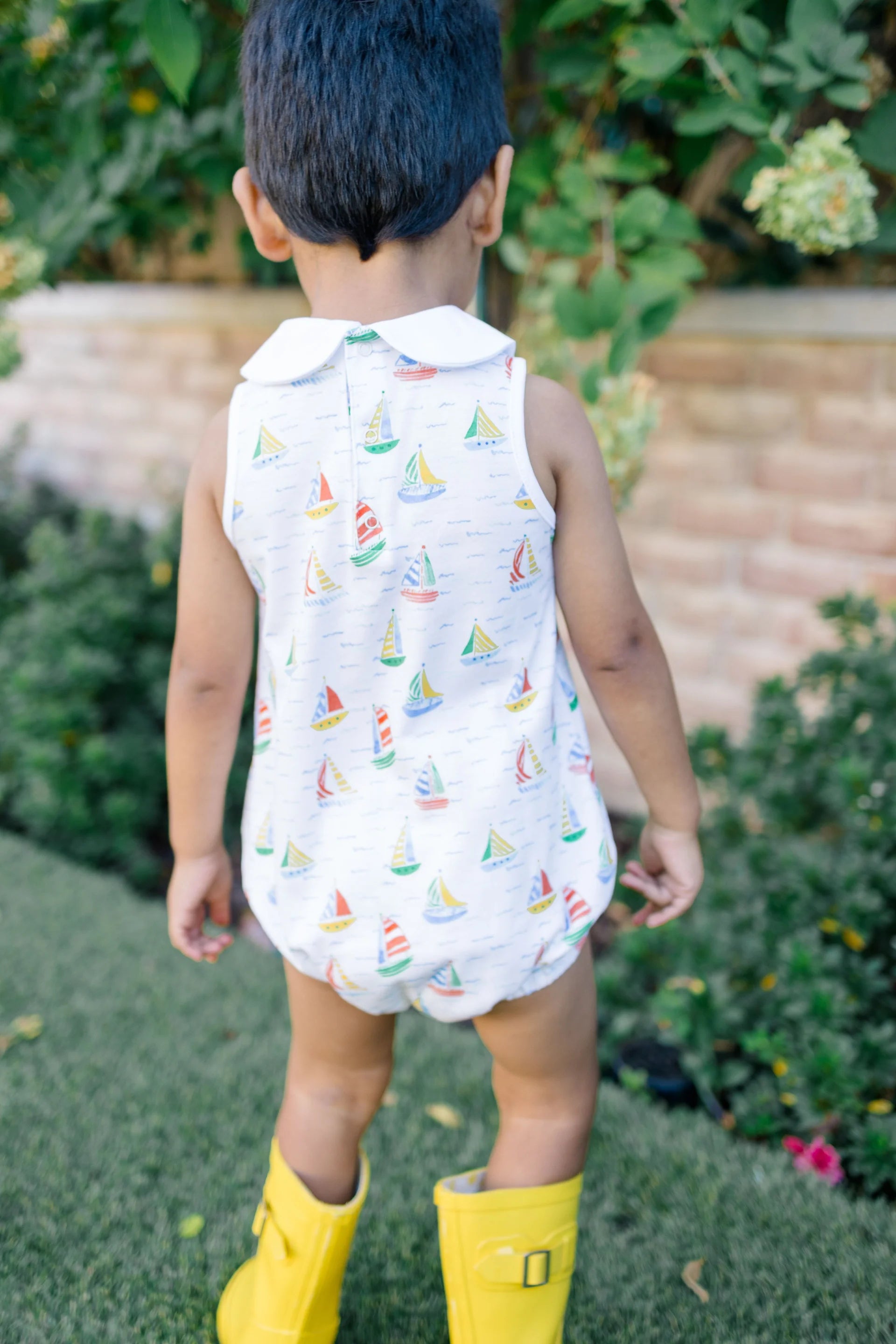 Child wearing a sleeveless romper with sailboat pattern and yellow rain boots outdoors.