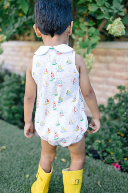 Child wearing a sleeveless romper with sailboat pattern and yellow rain boots outdoors.