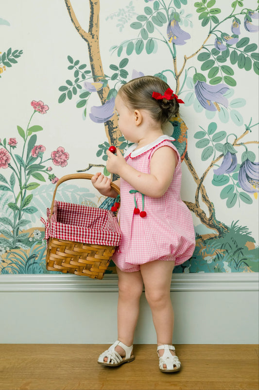 Child in a pink dress holding a picnic basket against a floral wallpapered wall.