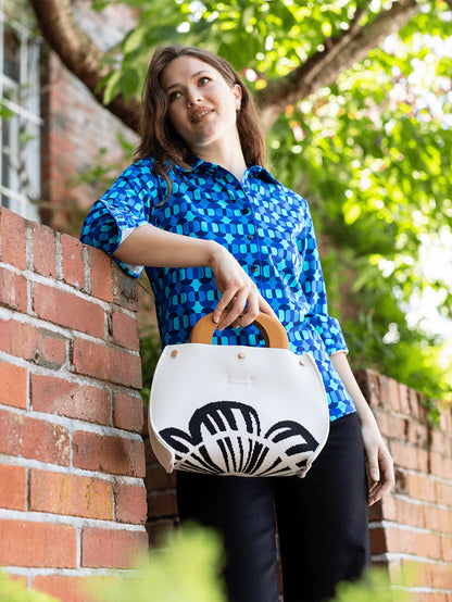 Woman holding a white bag with black design, standing outdoors near a brick wall and tree.