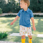 Child wearing a blue shirt, patterned shorts, and yellow rain boots outdoors.