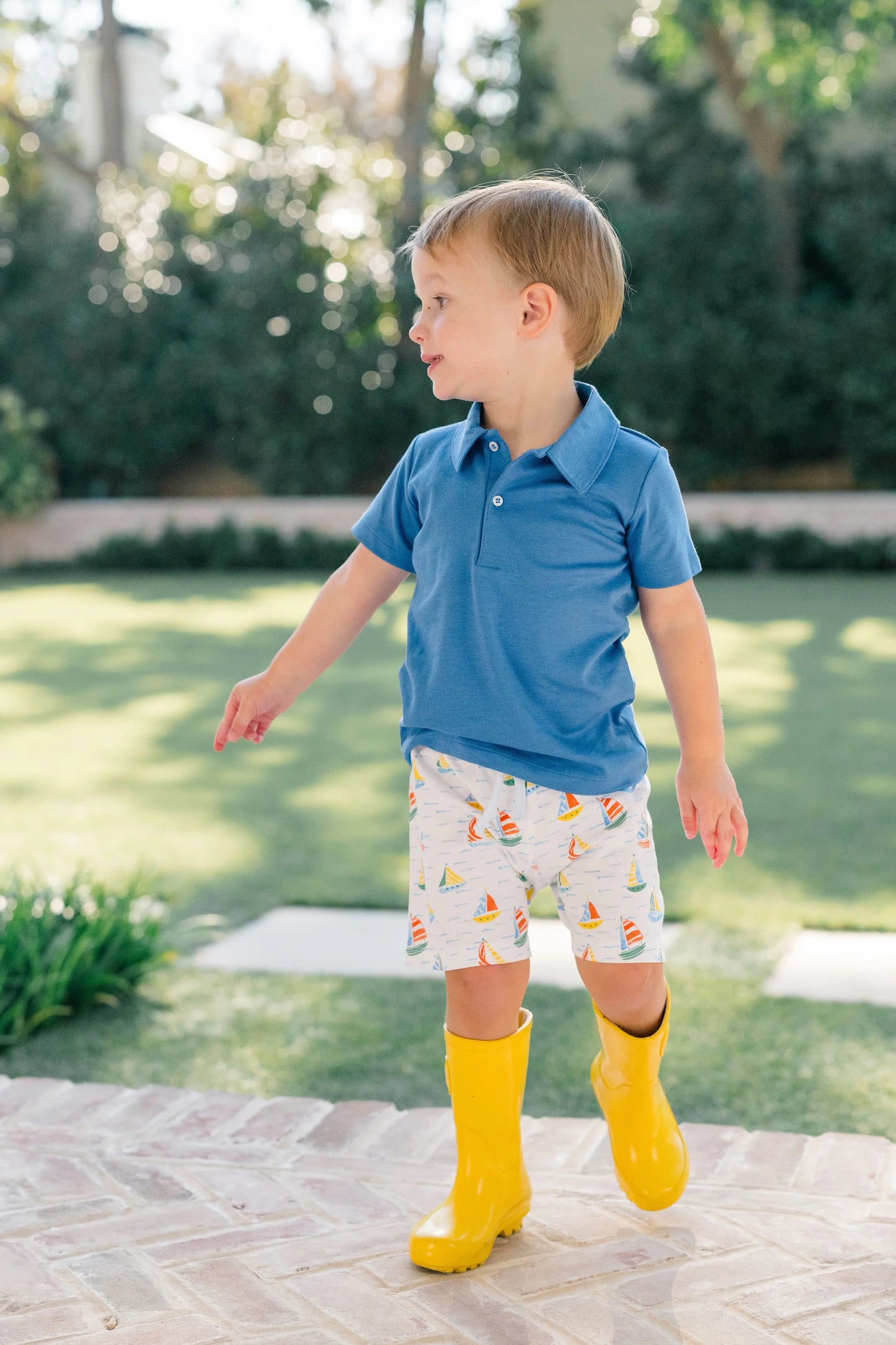 Child wearing a blue shirt, patterned shorts, and yellow rain boots outdoors.