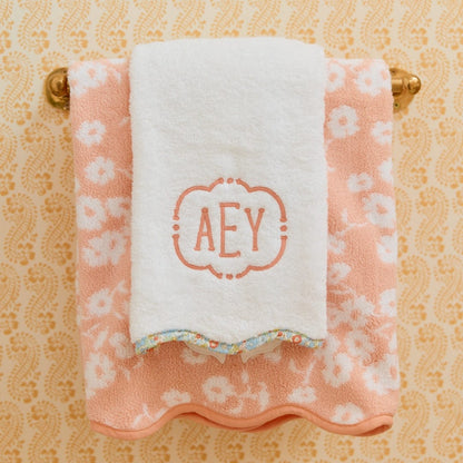 White towel with 'AEY' monogram on a pink floral towel, both hanging on a gold towel rack against a patterned wall.