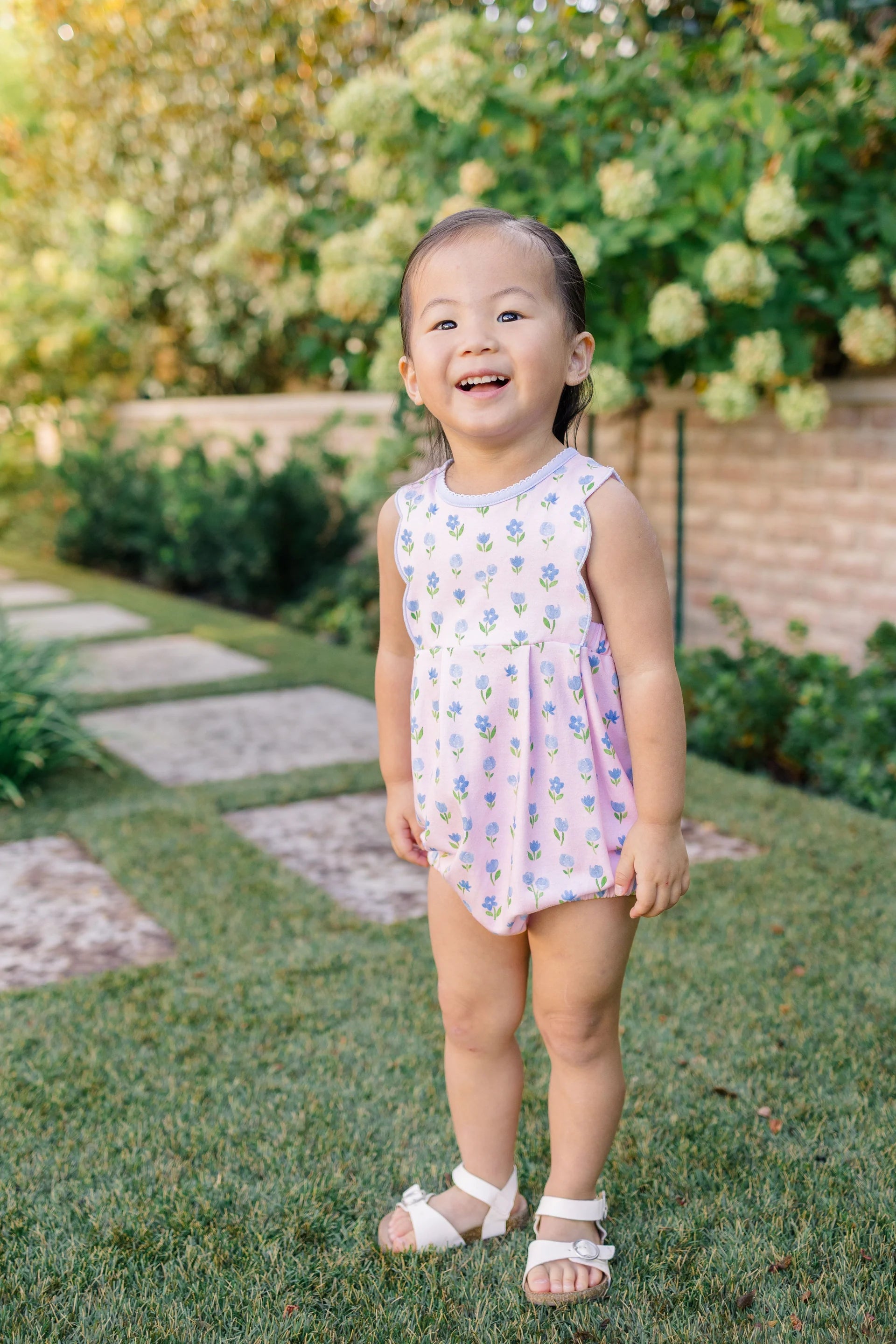 Child in a floral dress standing on grass with greenery in the background