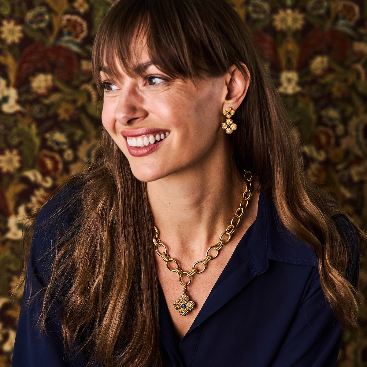 Woman wearing a gold necklace and earrings against a patterned wall.