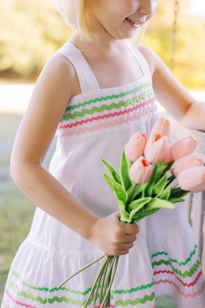 A white Sundress with multicolored ric rac trim and a bow at the back neck, designed for children.