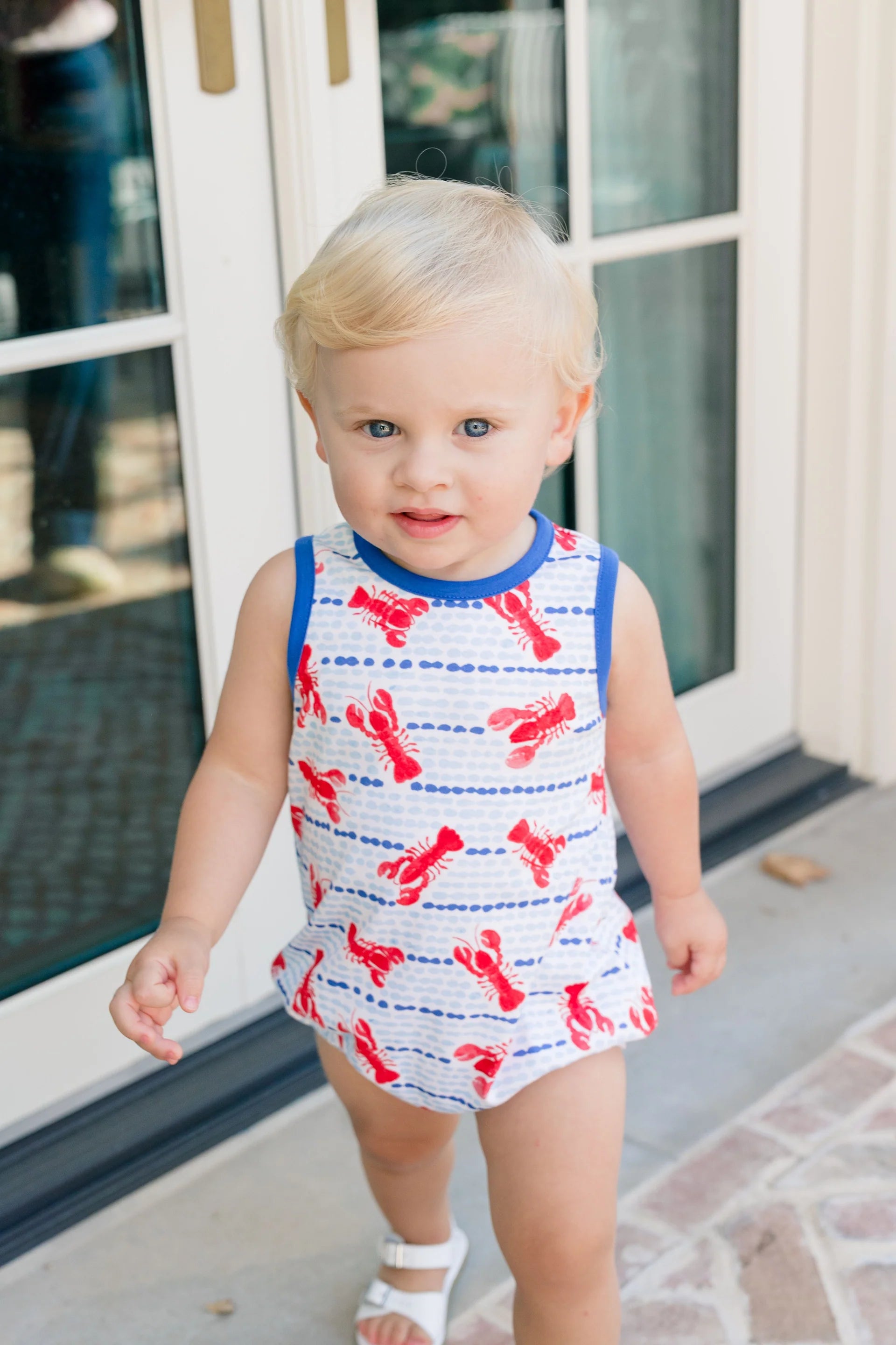 Child wearing a red and blue swimsuit with lobster pattern standing in front of a glass door.