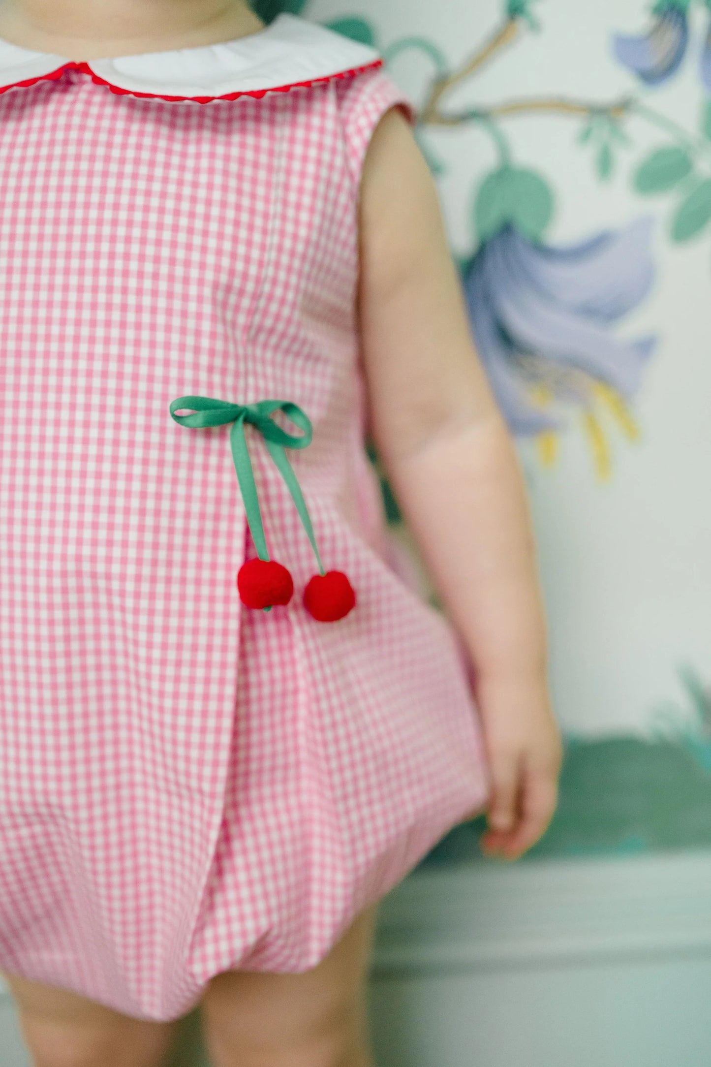 Child wearing a pink checkered romper with cherry details against a floral background