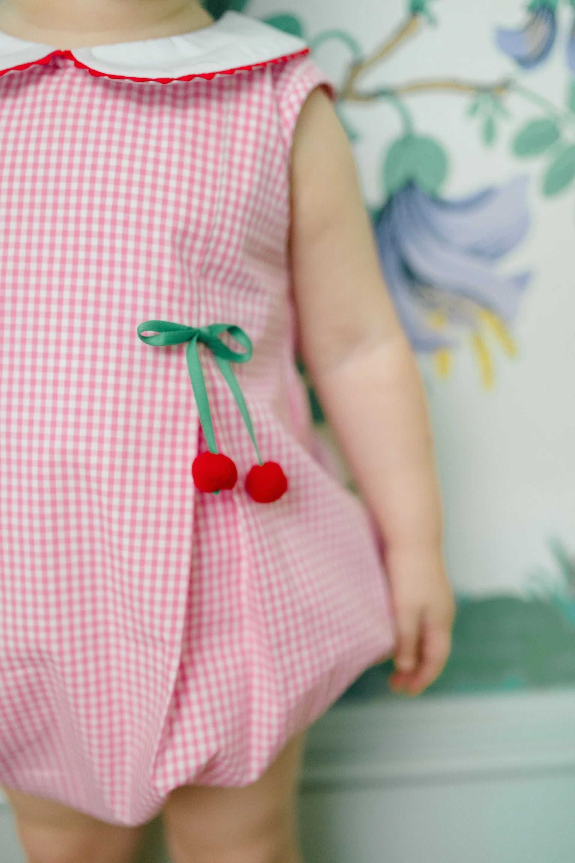 Child wearing a pink checkered romper with cherry details against a floral background