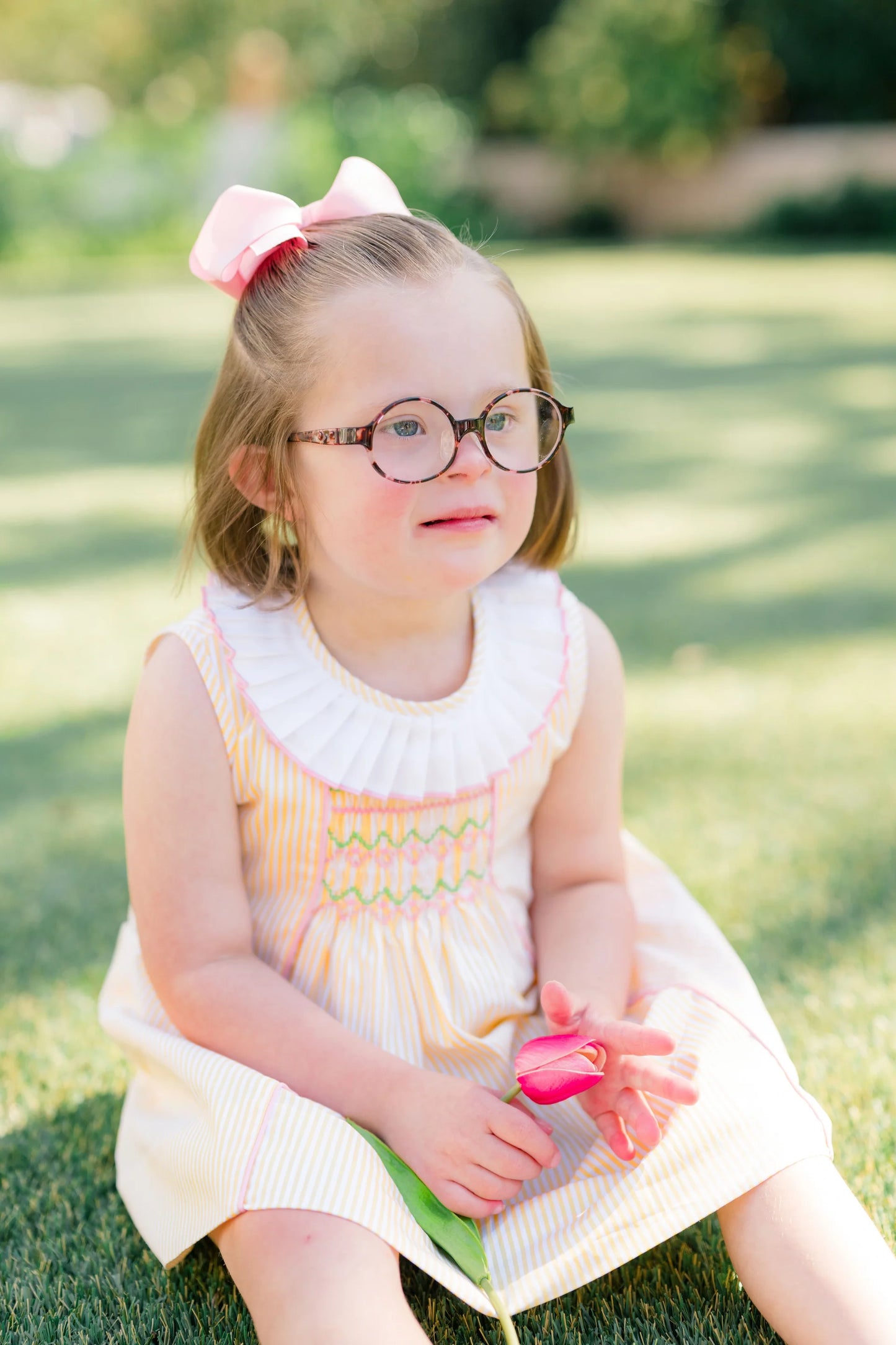 Young girl with glasses and a pink bow sitting on grass holding a flower