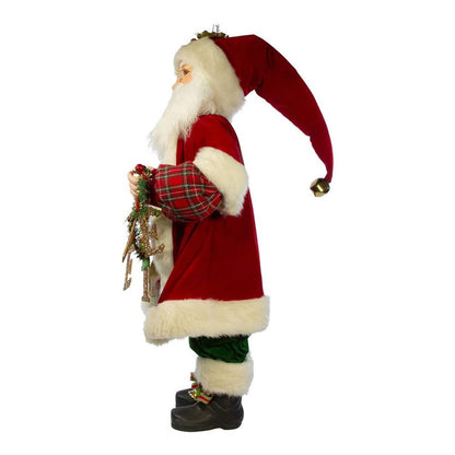 A Santa Claus Christmas decoration figure shown in profile against a plain white background. The figure wears a red coat with white faux-fur trim, a long red hat with a gold bell, green pants, and black boots, and holds a small holiday wreath.