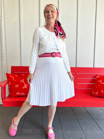 Woman in a white outfit with a red belt and pink shoes standing on a wooden floor.