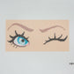 Cross-stitched design of an eye with eyelashes on a beige background, featuring the brand 'Milli Tomas' logo.