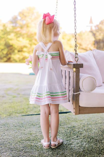 A white Sundress with multicolored ric rac trim and a bow at the back neck, designed for children.