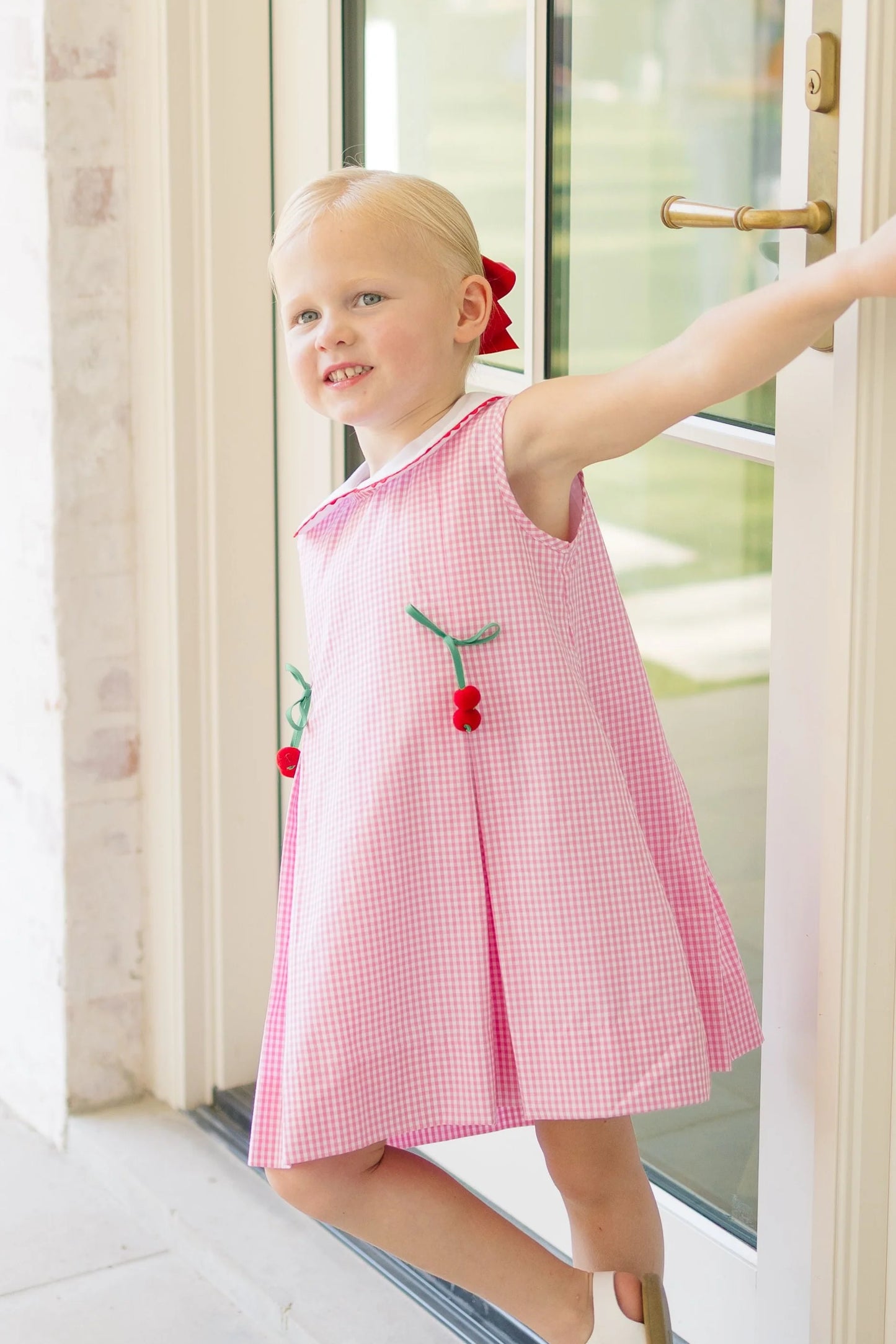 Young girl in a pink dress with red accents standing by a door.