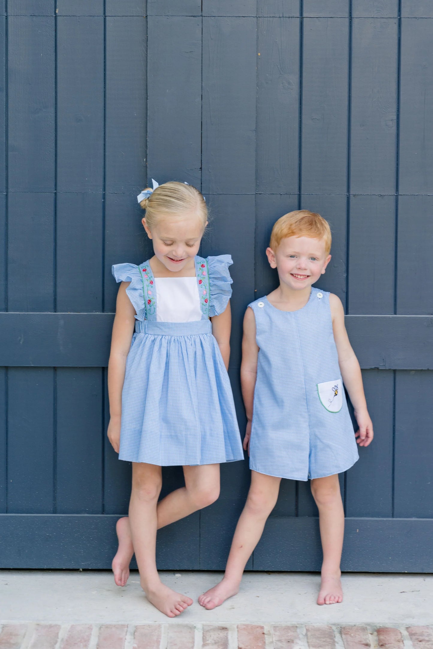 Two children in matching blue dresses standing against a dark blue wooden wall.