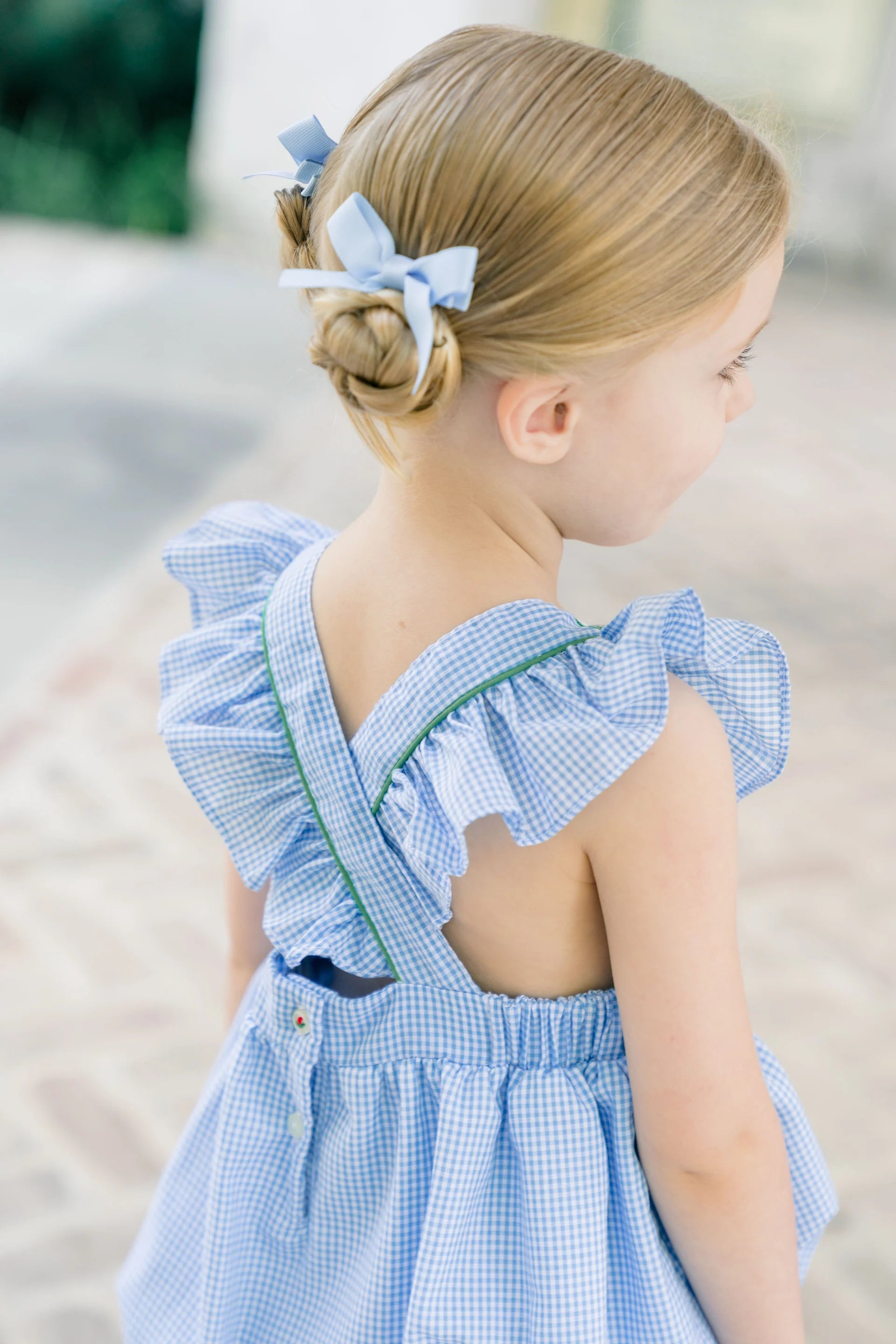 Young girl wearing a blue checkered dress with ruffled sleeves and a bow in her hair.
