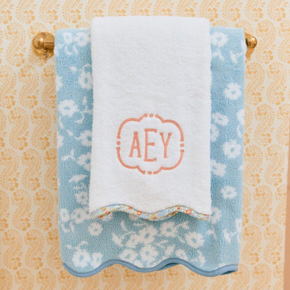 White towel with 'AEY' monogram on a floral-patterned blue towel, hanging on a gold towel rack.